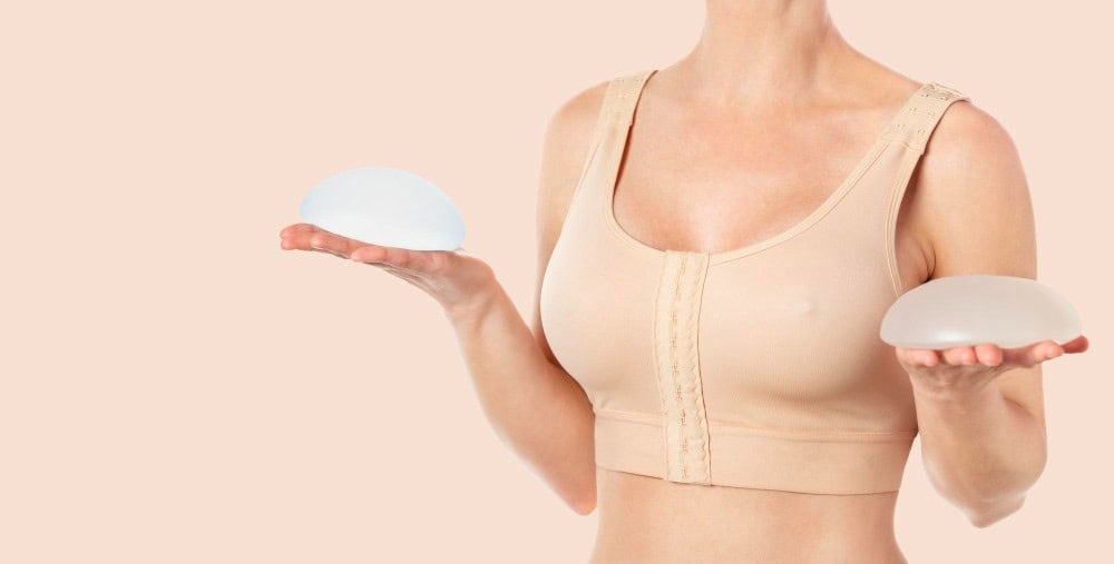 Woman in a post-surgery bra holding breast implants during breast augmentation recovery in Albania